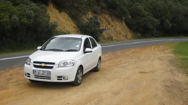 A white car is parked on a sandy pullout beside a winding road.