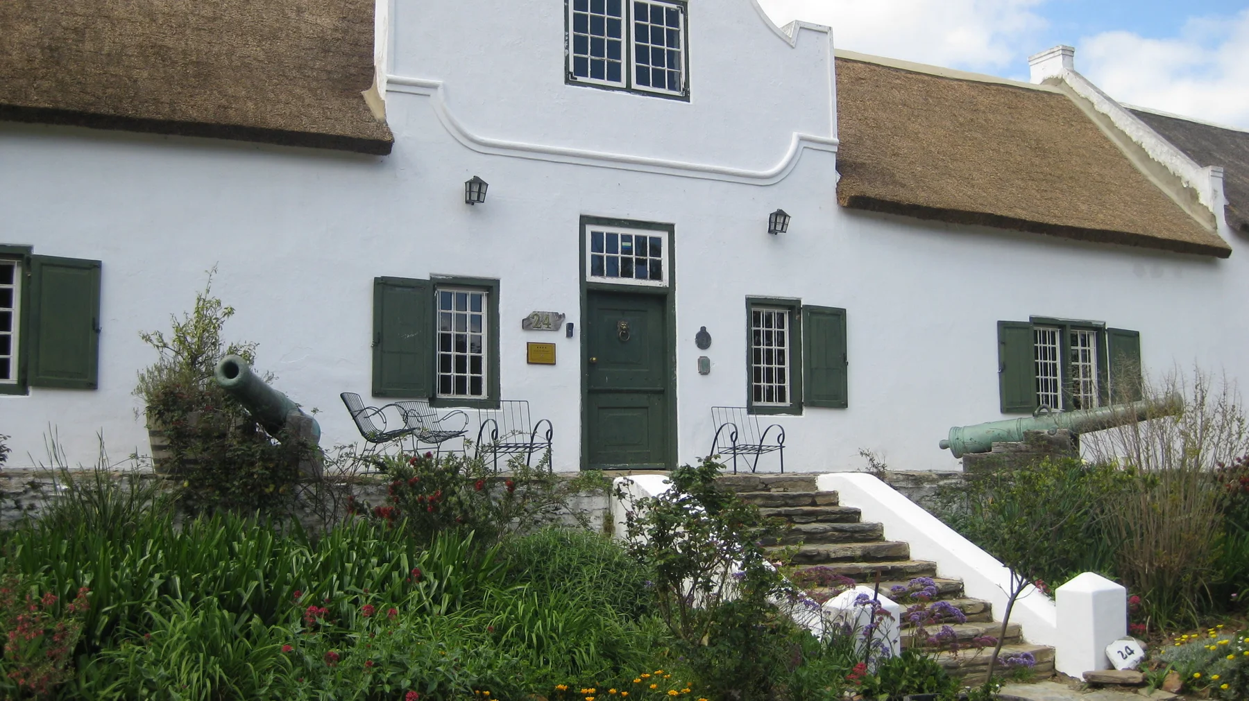 Green shutters and a dark door punctuate a white thatched house behind a garden.