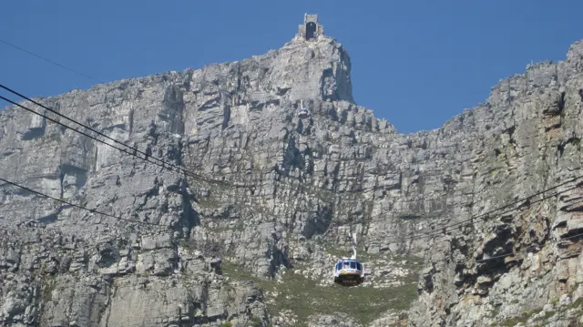 A cable car hangs beneath cables against a sheer rock face.