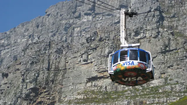 A rounded cable car passes in front of a layered cliff face.