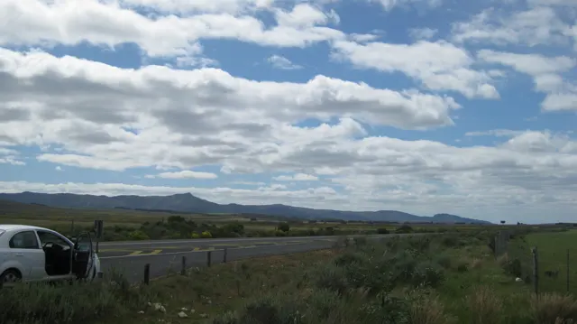 A parked car sits beside open fields beneath a wide cloud-filled sky.