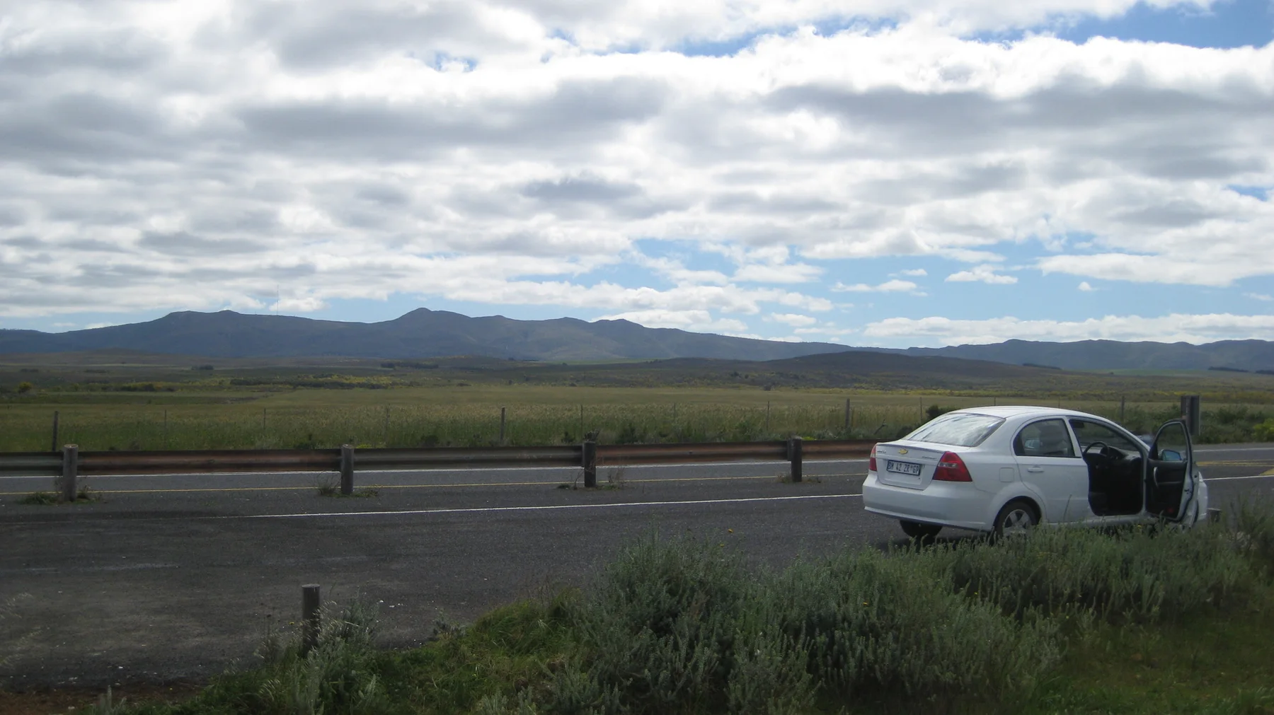 A white car with an open door stands beside a road through open fields.