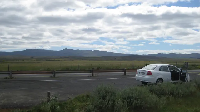 A white car with an open door stands beside a road through open fields.