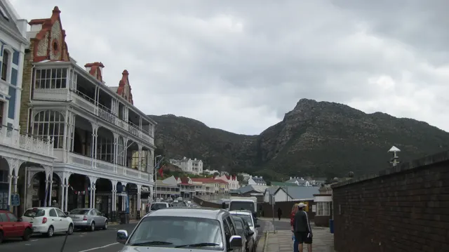 Ornate buildings and parked cars line a street beneath a rocky mountain.