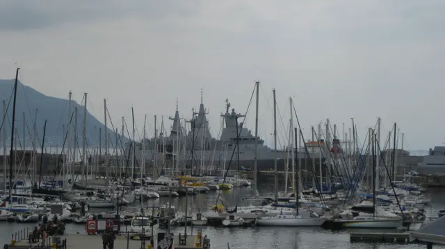 Dense sailboat masts rise before waterfront buildings and distant mountains.