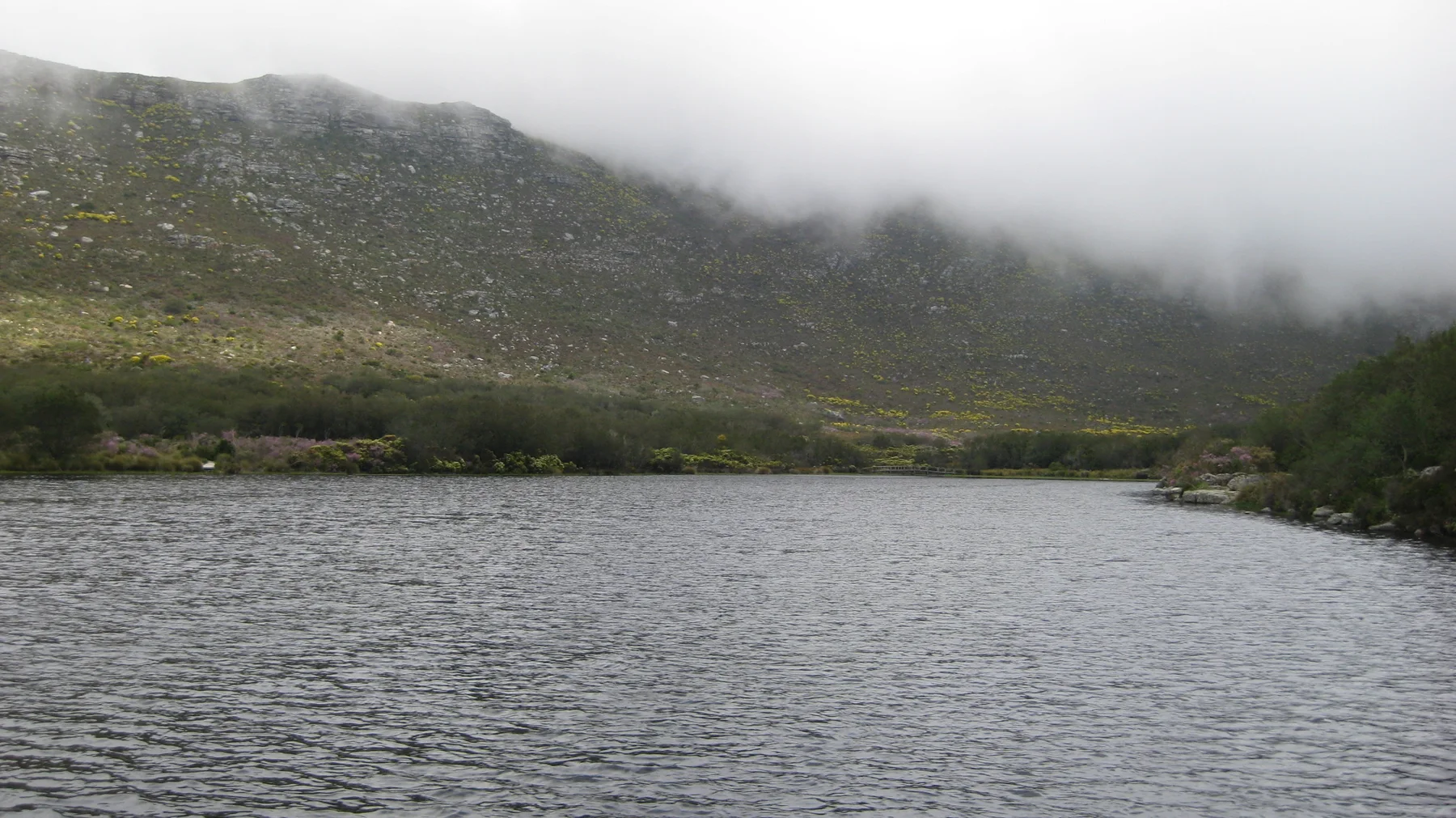 Rippling reservoir water lies below a hillside covered by low cloud.