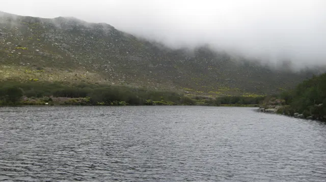 Rippling reservoir water lies below a hillside covered by low cloud.
