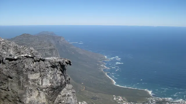 A rough rock ledge overlooks a long mountain coastline and deep blue sea.