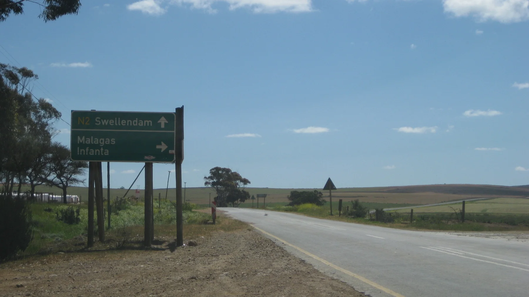 A green direction sign stands beside a quiet road through open land.