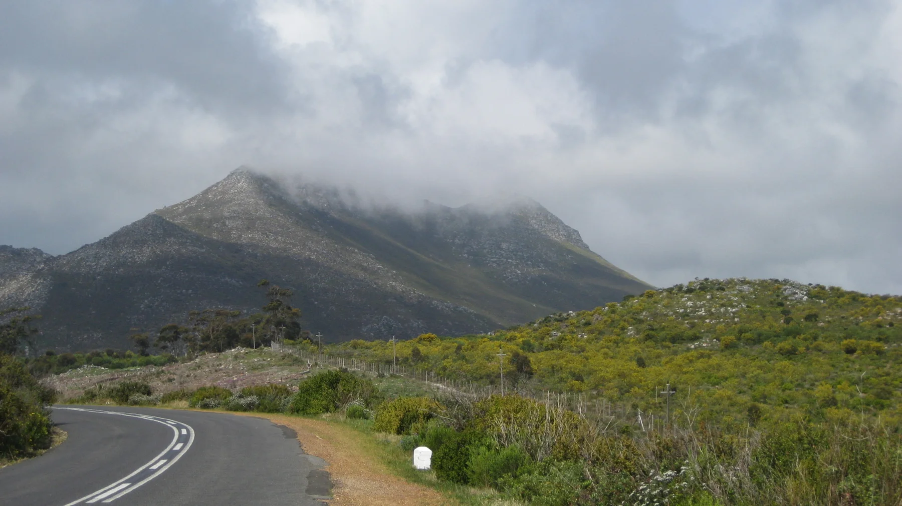 A road curves through scrub toward a mountain topped with cloud.