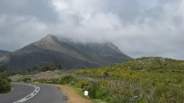 A road curves through scrub toward a mountain topped with cloud.