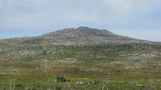 A low stony hill rises above sparse green scrub under clear sky.