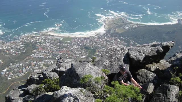 A person sits among rocks high above coastal buildings and blue water.