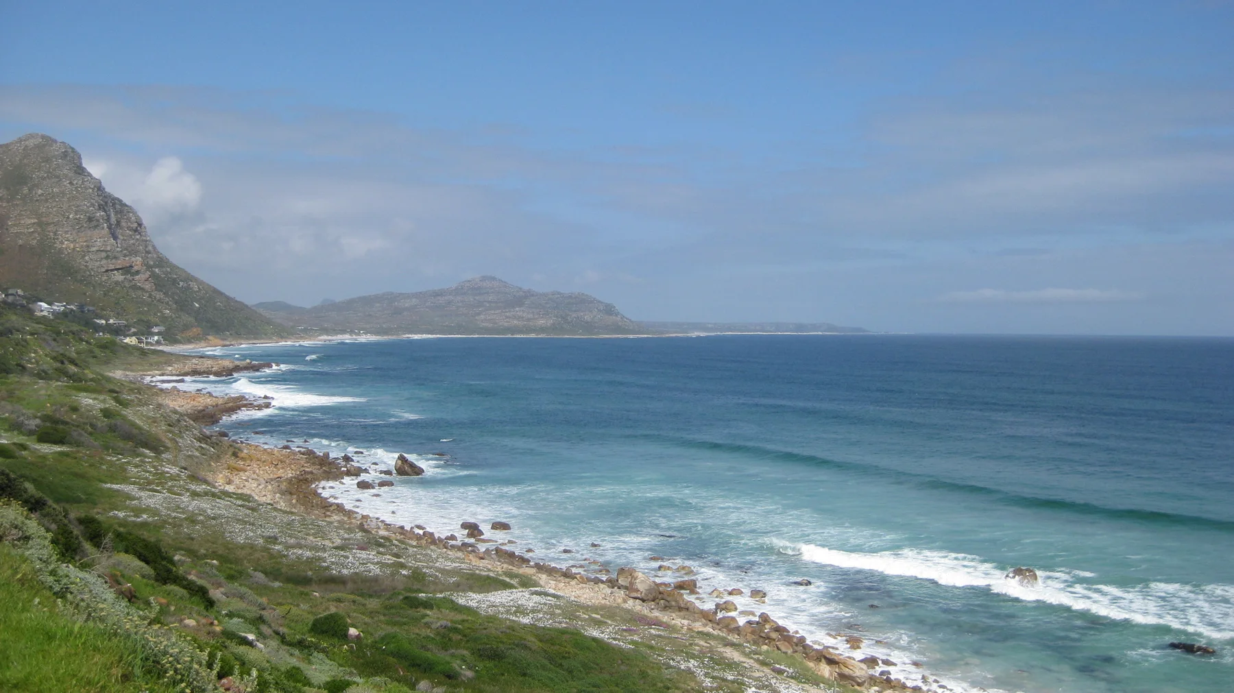 Blue waves break along a rocky coast below steep mountains.