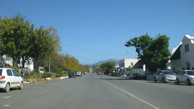 A broad street passes pale buildings and parked cars beneath blue sky.