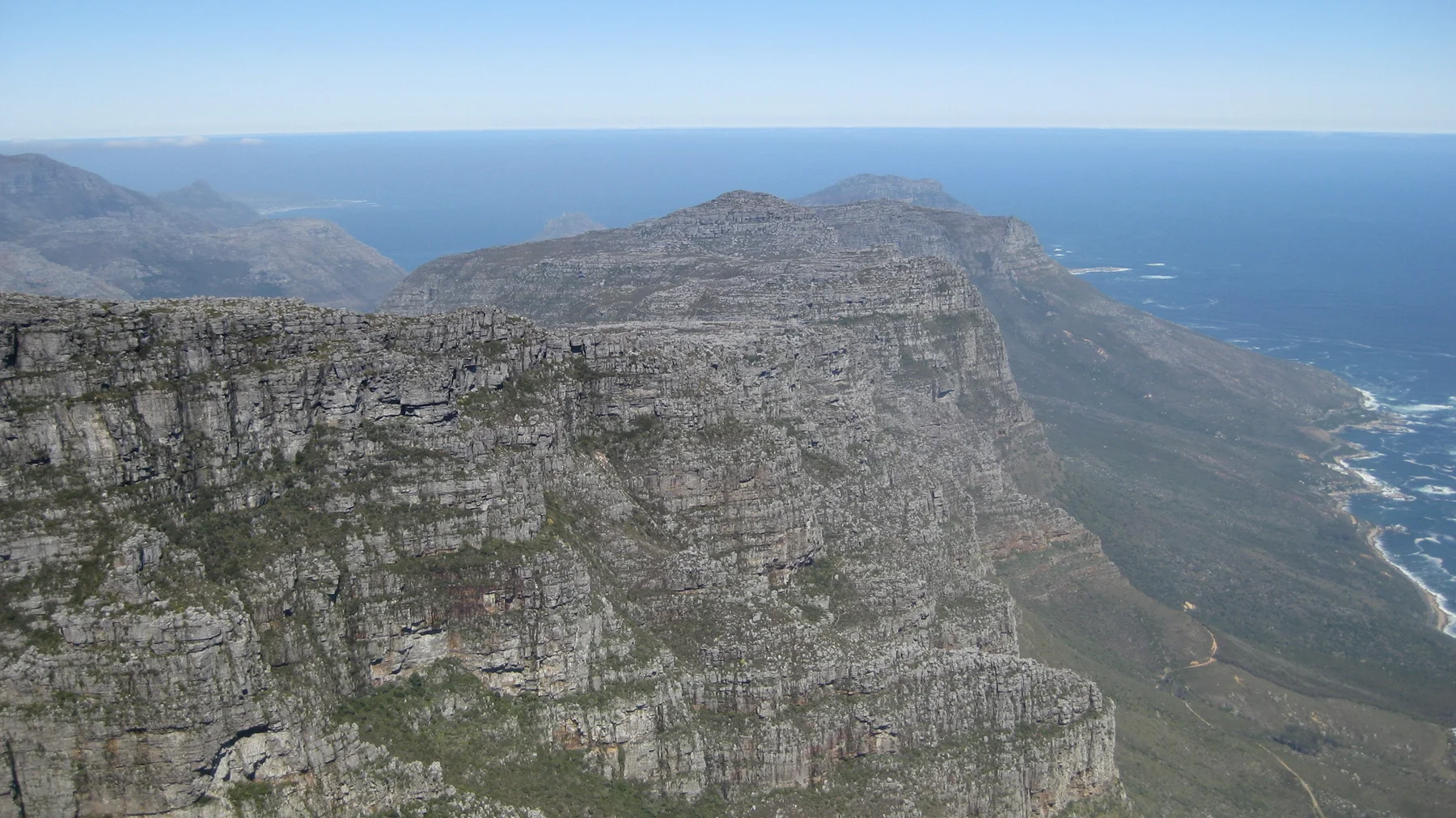 A sheer cliff overlooks a mountain coastline and deep blue ocean.