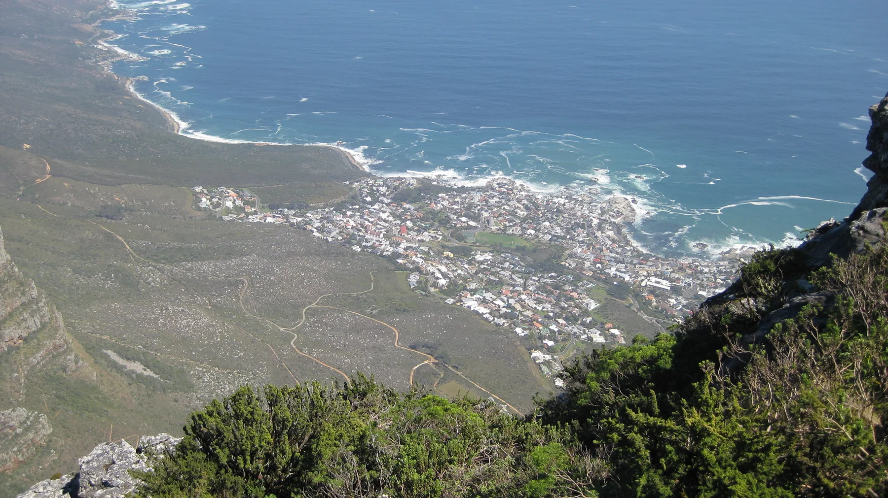 Coastal buildings and white surf lie far below a steep green slope.