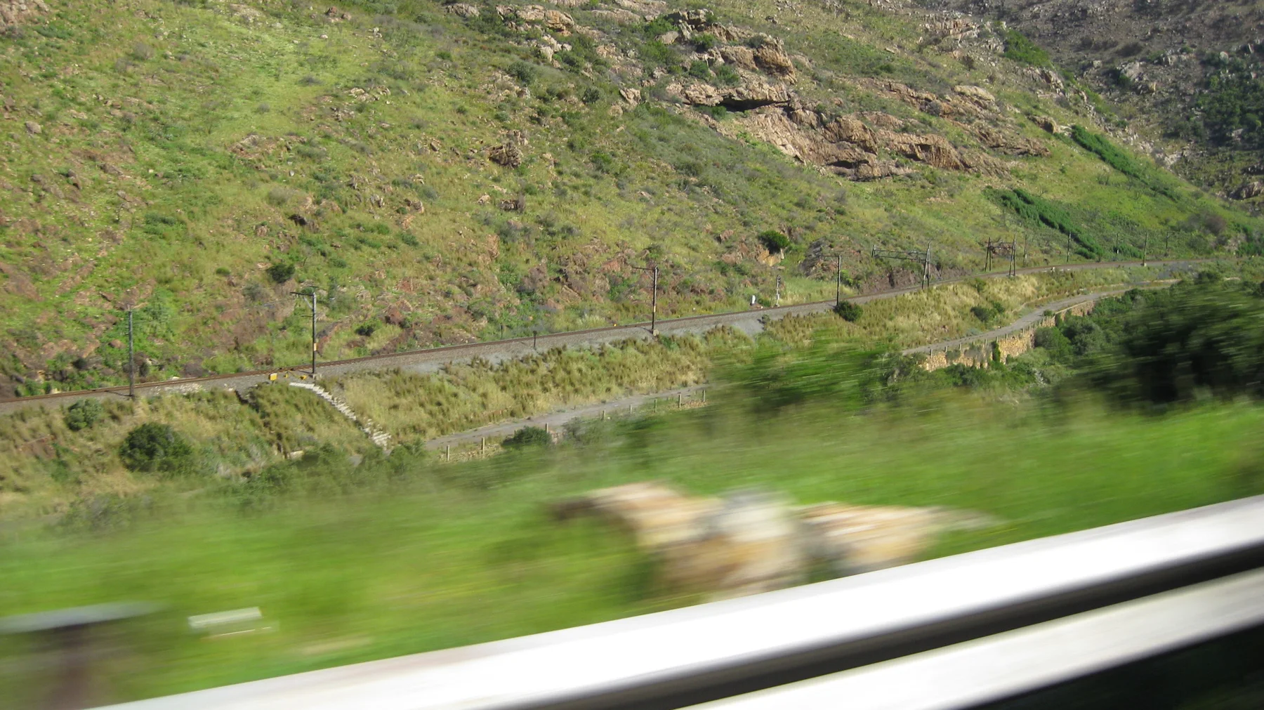 A road cuts across a green rocky slope beyond a blurred guardrail.