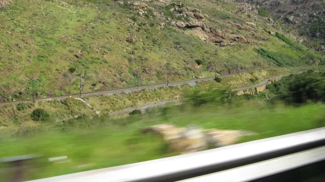 A road cuts across a green rocky slope beyond a blurred guardrail.
