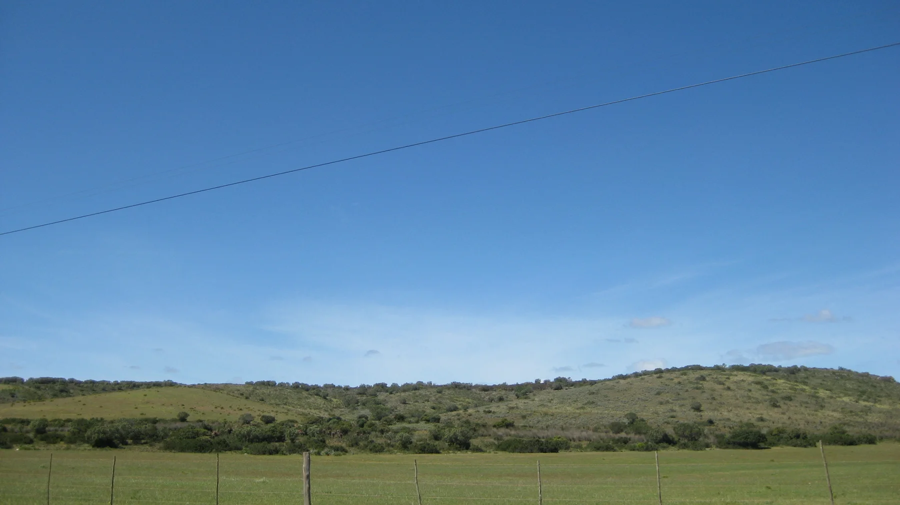 A utility line crosses blue sky above fenced fields and low hills.