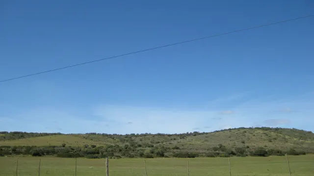 A utility line crosses blue sky above fenced fields and low hills.