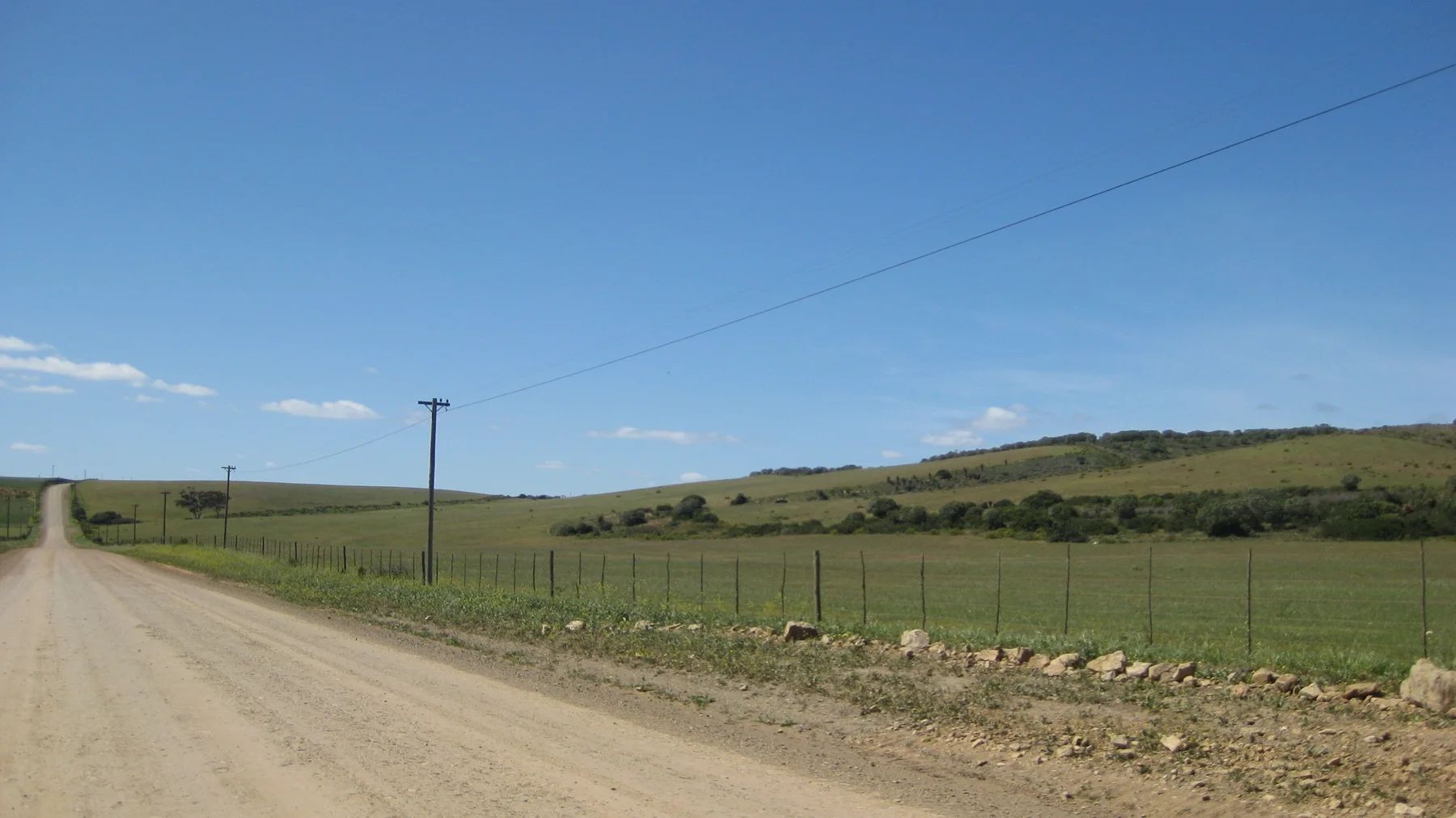 A dirt road passes fenced fields and utility poles under clear sky.