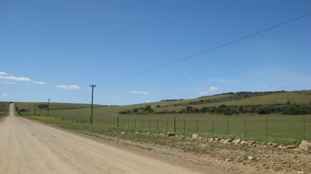 A dirt road passes fenced fields and utility poles under clear sky.