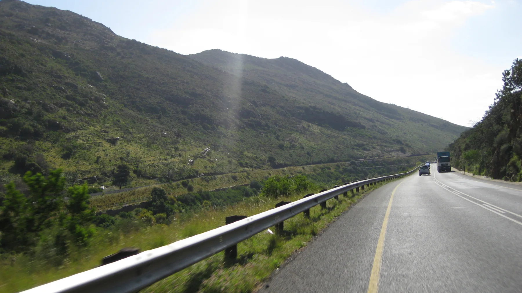 A road and metal guardrail follow the base of a steep green mountain.
