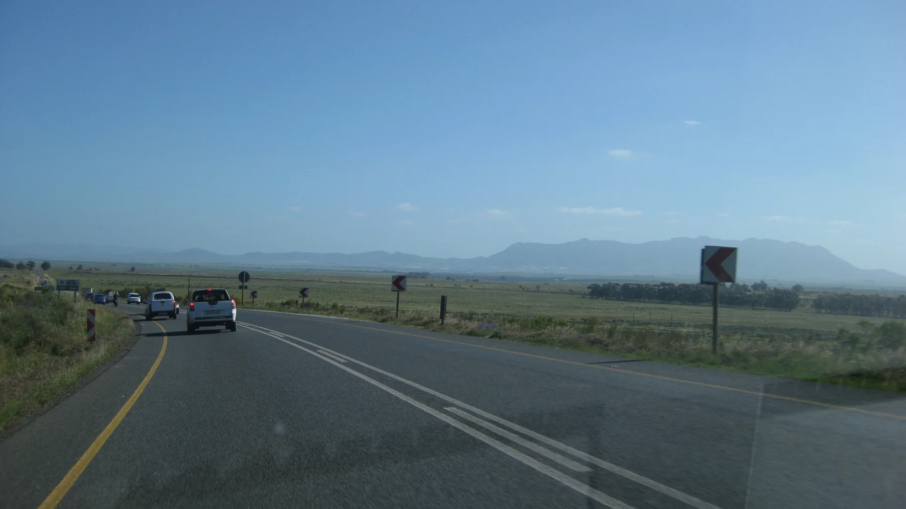 Cars follow a bend through open fields beneath a distant mountain silhouette.