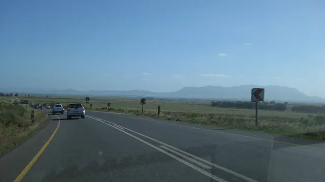 Cars follow a bend through open fields beneath a distant mountain silhouette.