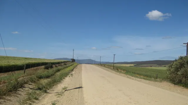 A straight dirt road crosses rolling farmland beneath a bright blue sky.