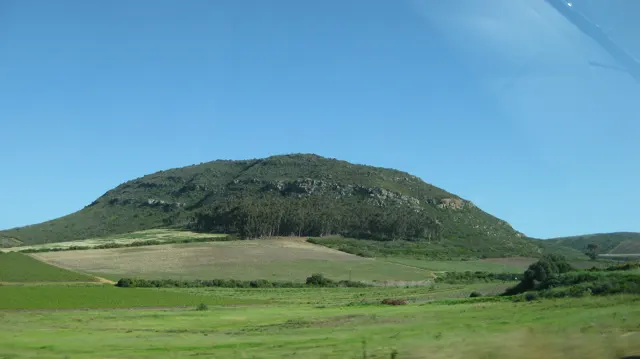 A rounded green hill rises above patchwork fields under clear sky.
