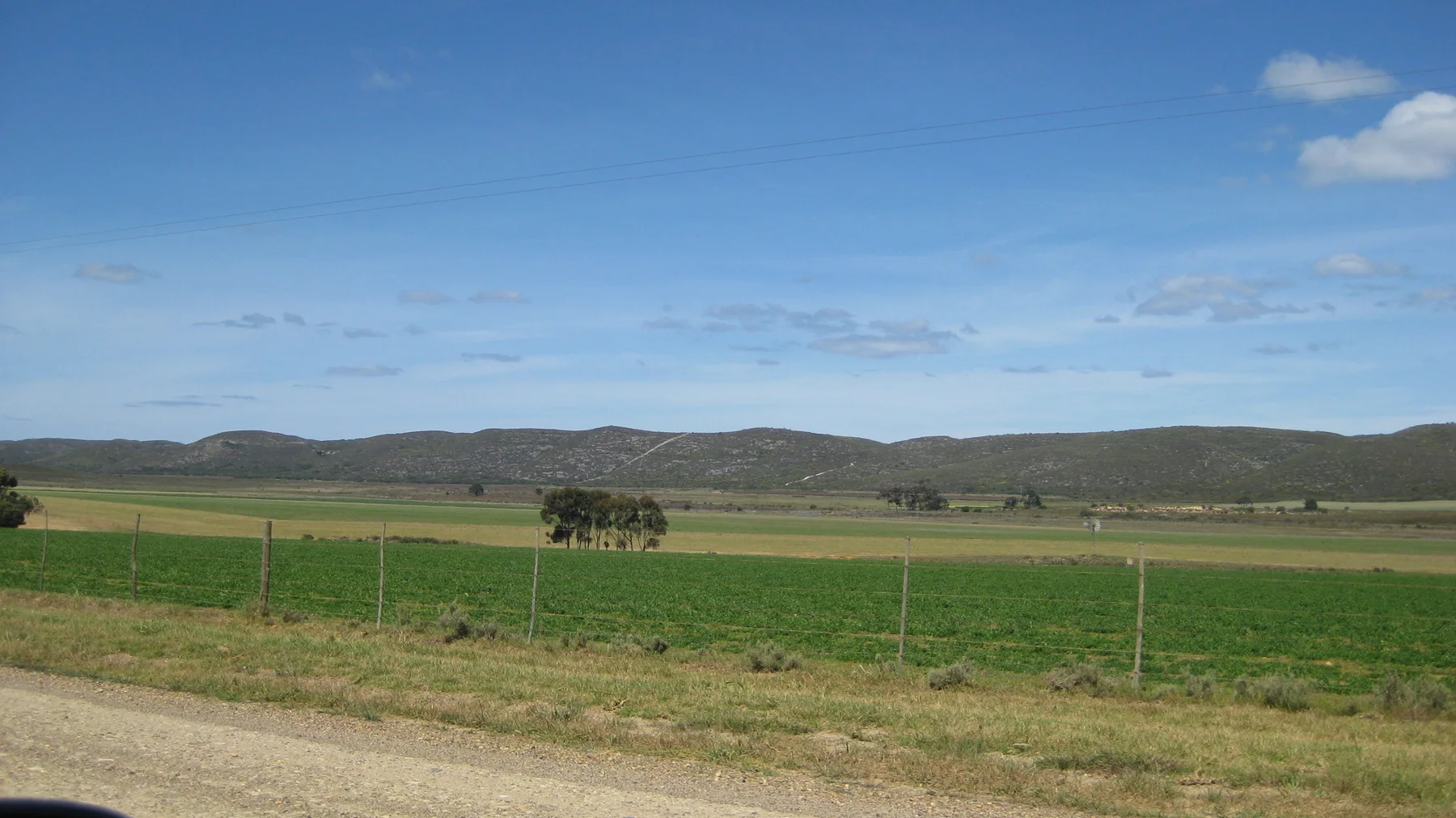Fenced green fields stretch beyond a gravel roadside toward low hills.