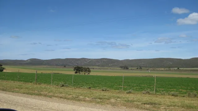 Fenced green fields stretch beyond a gravel roadside toward low hills.