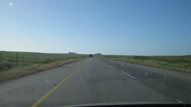A straight road crosses flat fields under cloudless blue sky.