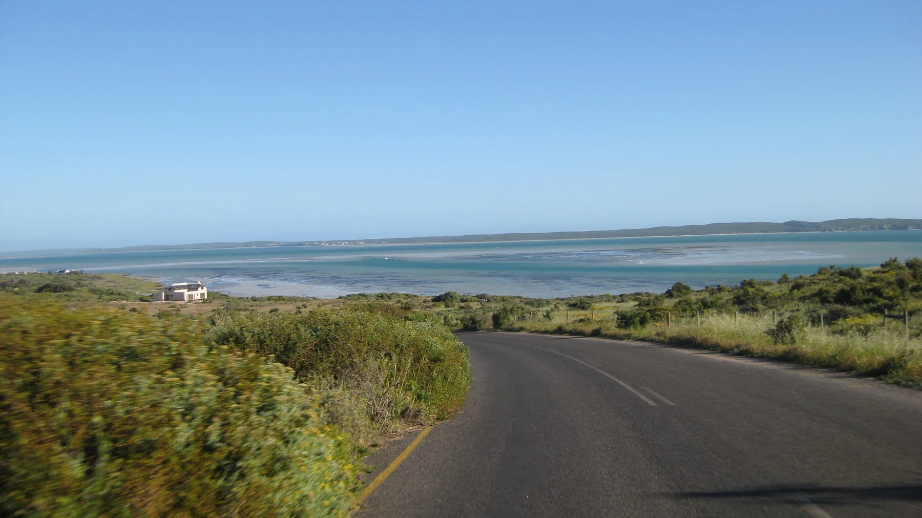 A road curves downhill through shrubs toward broad blue water.
