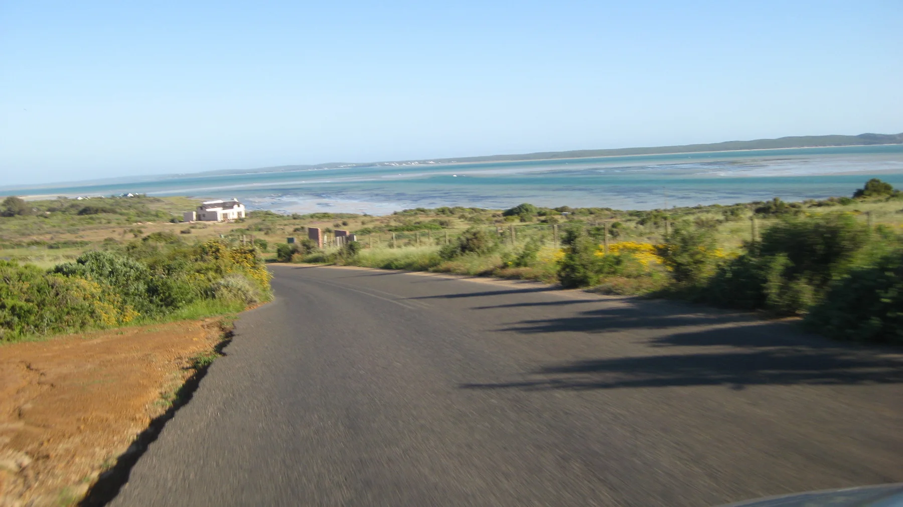 A narrow road descends through low scrub toward pale blue water.