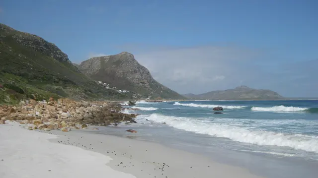 White surf rolls past beach rocks below a steep mountain.