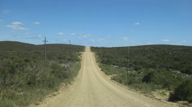 A straight gravel road climbs gently between shrubs and utility poles.