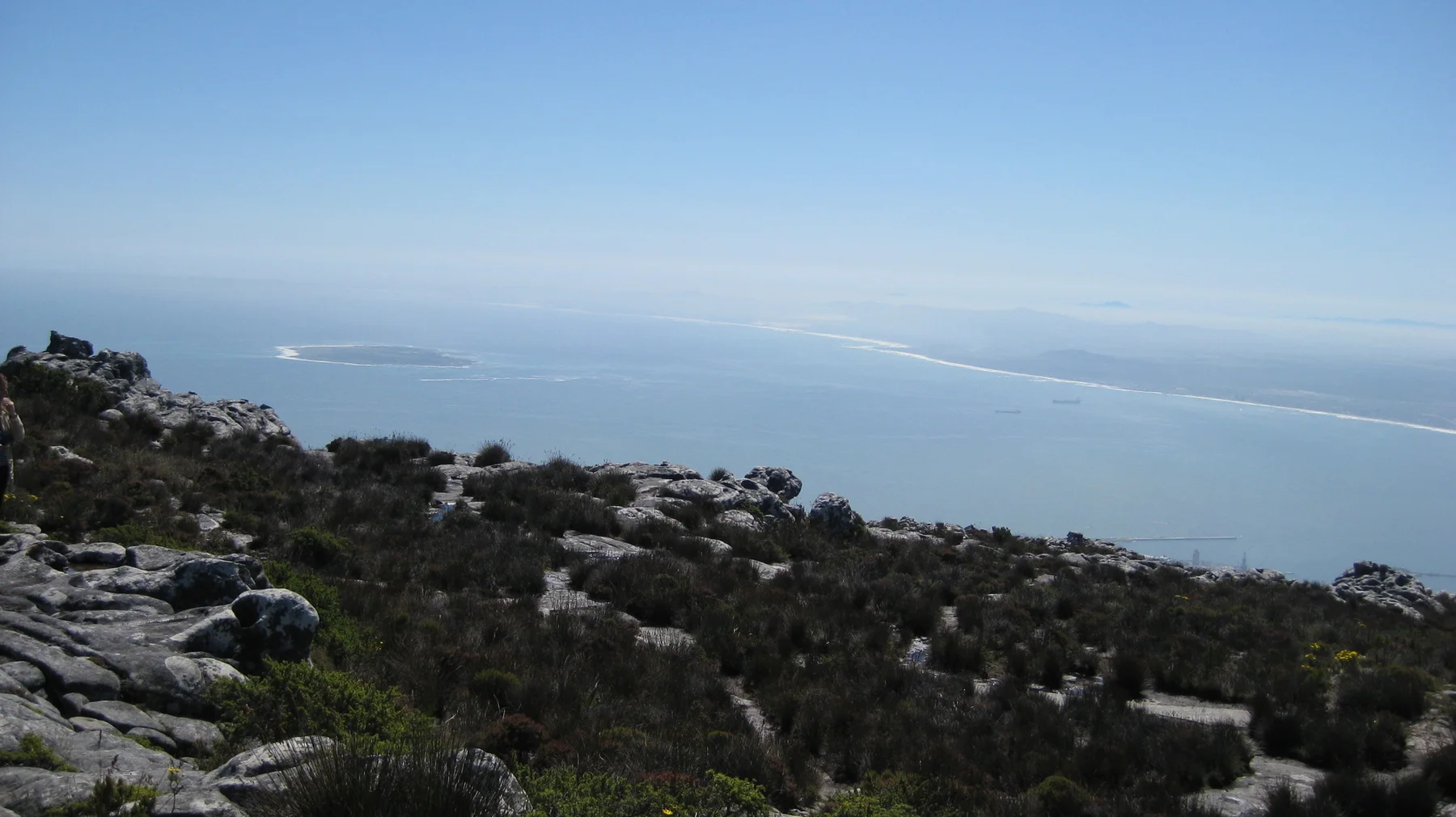 Rocky foreground slopes overlook a hazy coastline and pale blue water.