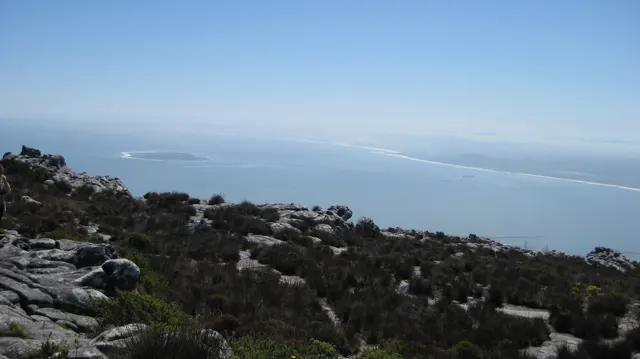 Rocky foreground slopes overlook a hazy coastline and pale blue water.