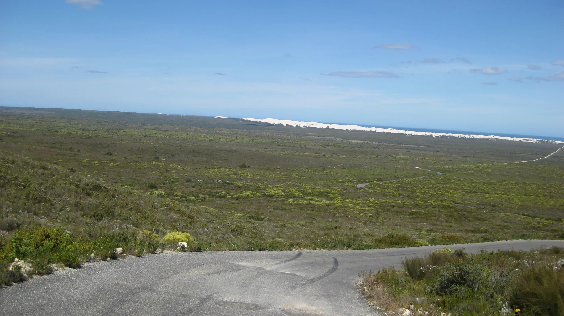 A paved road crosses low scrub toward a distant white coastal strip.