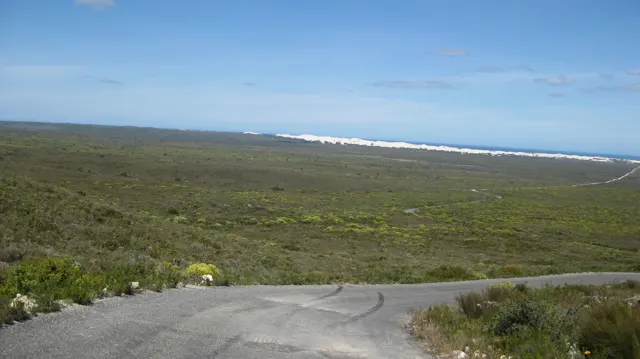 A paved road crosses low scrub toward a distant white coastal strip.