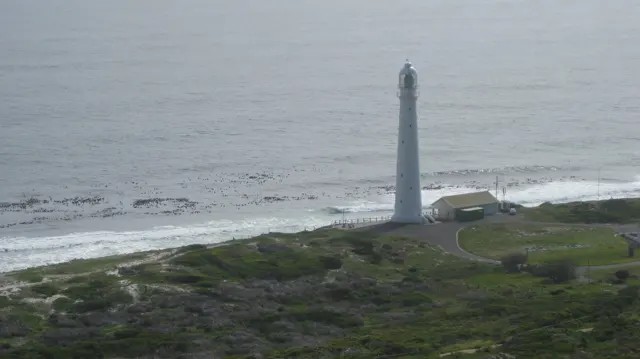 A tall pale lighthouse stands beside a narrow beach and gray sea.