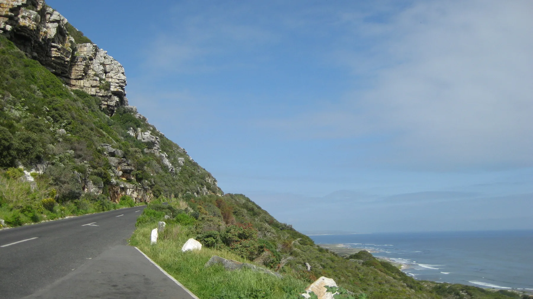 A paved road bends below rock and greenery above the sea.