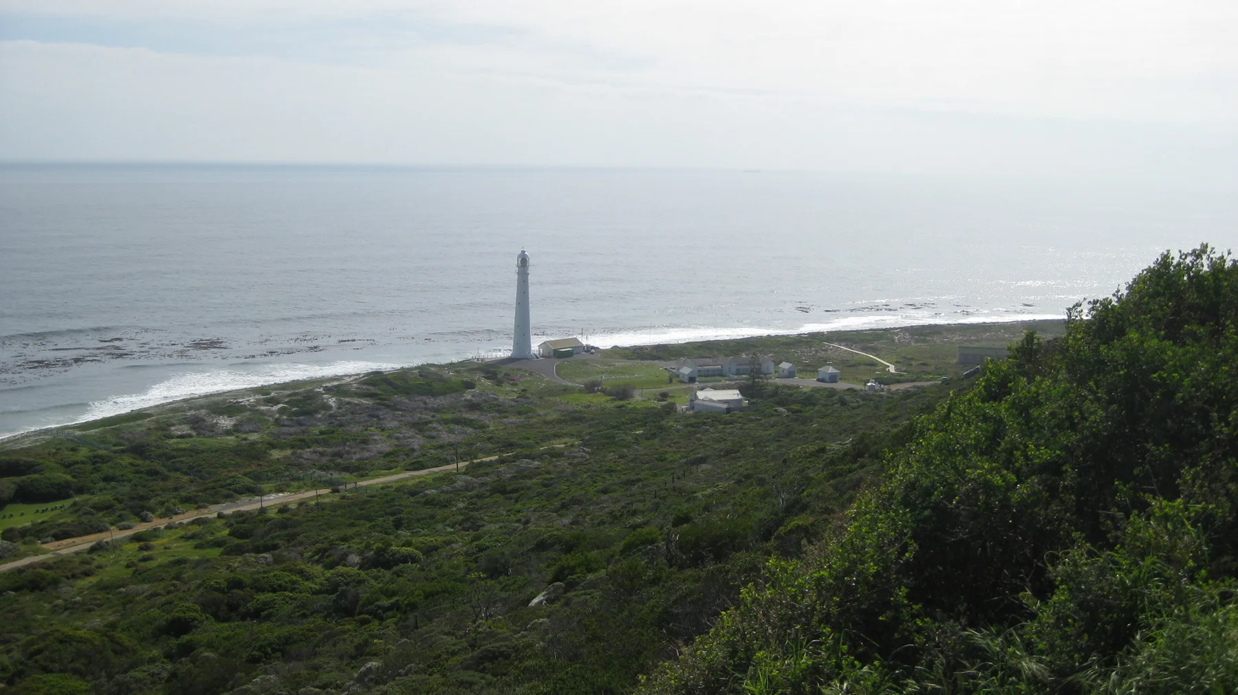 A distant slender lighthouse rises beside white surf and green coastal land.