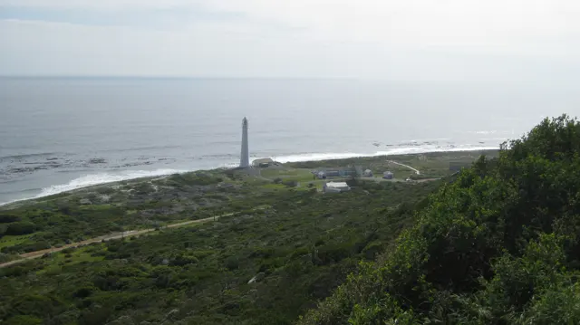A distant slender lighthouse rises beside white surf and green coastal land.