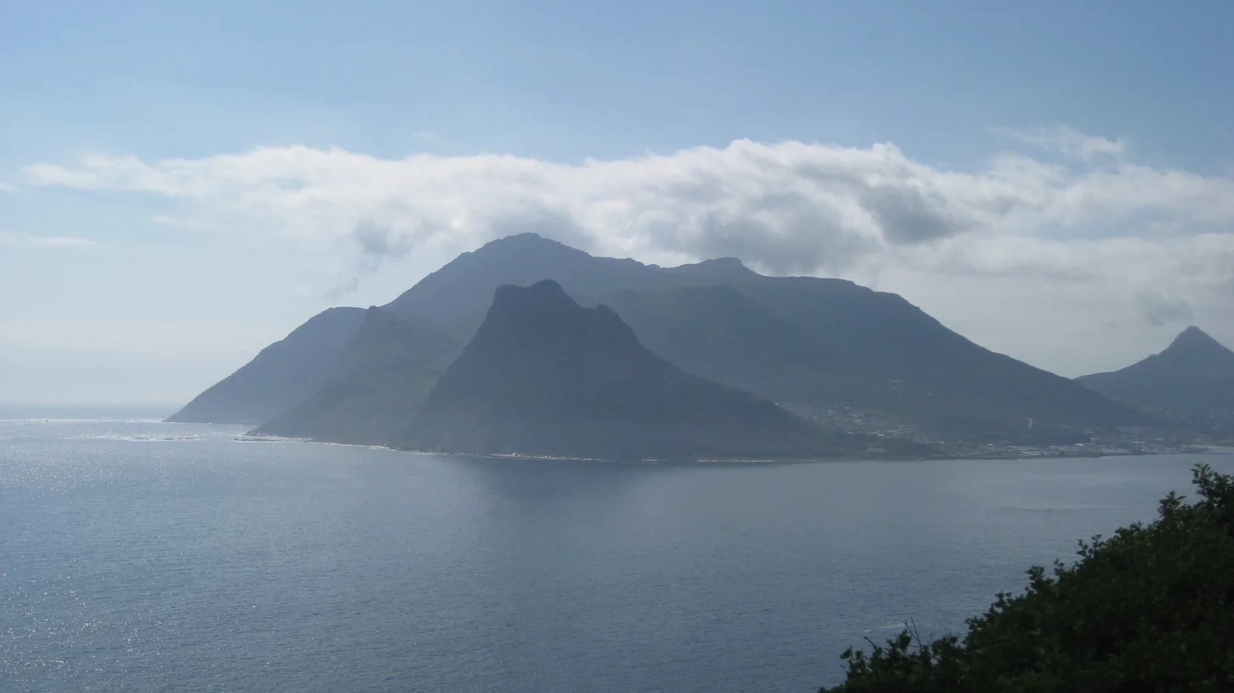 A dark mountain mass rises across calm bay water beneath white clouds.