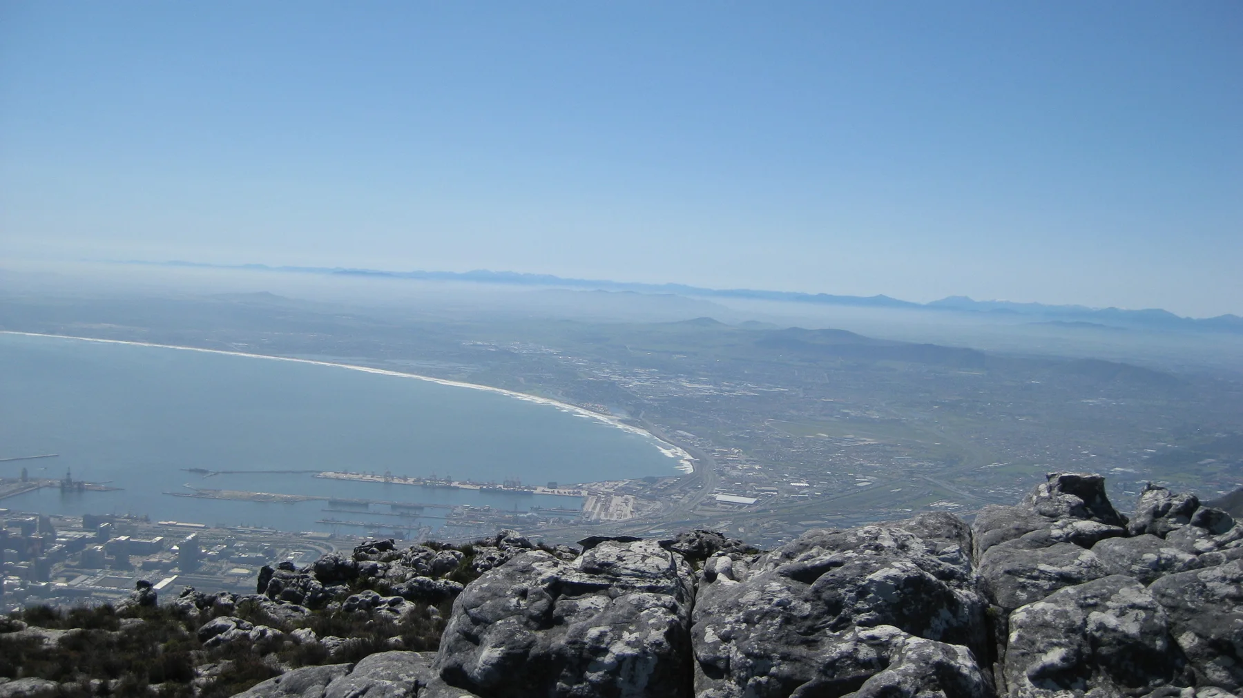 A sweeping bay curves beyond summit rocks and a hazy city.