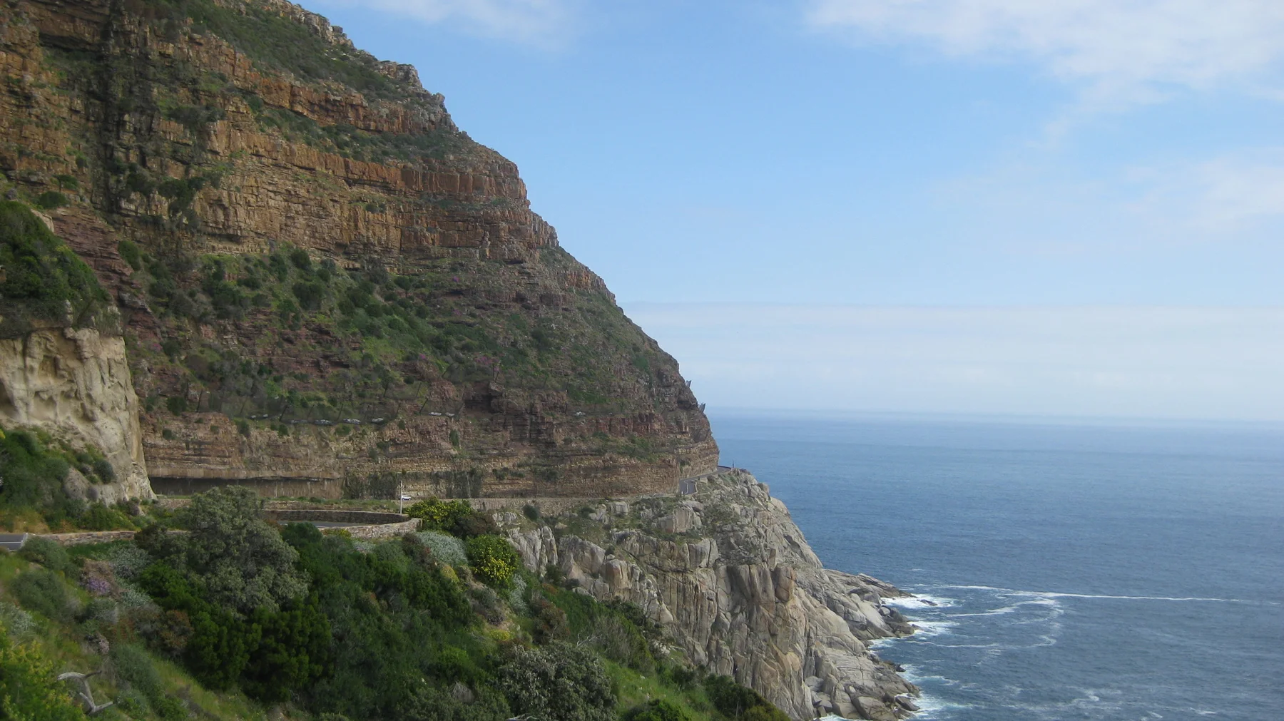 A narrow road follows a layered coastal cliff above blue sea.